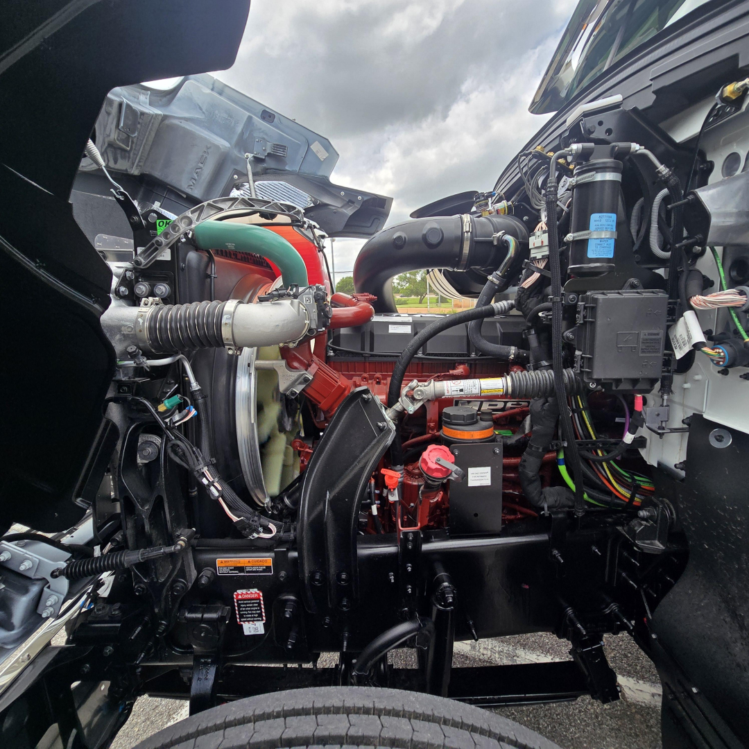 Truck engine bay view of a 2027 Mack PI64T showing engine components and hoses.