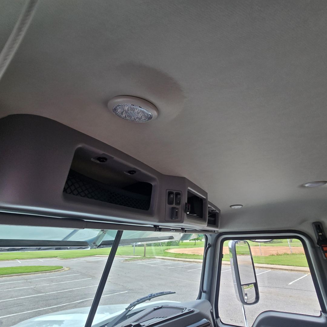 Interior cabin roof area of a 2027 Mack PI64T truck showing overhead storage cubby and dome light, with windshield and side mirror visible.