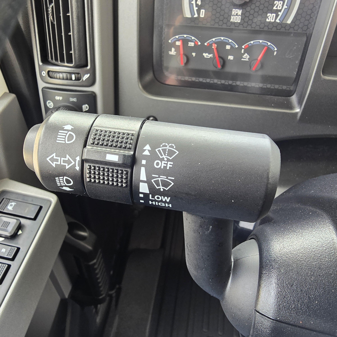 Truck steering column stalk with controls showing turn signals, wipers, and headlight/indicator icons; dashboard gauges visible in background.