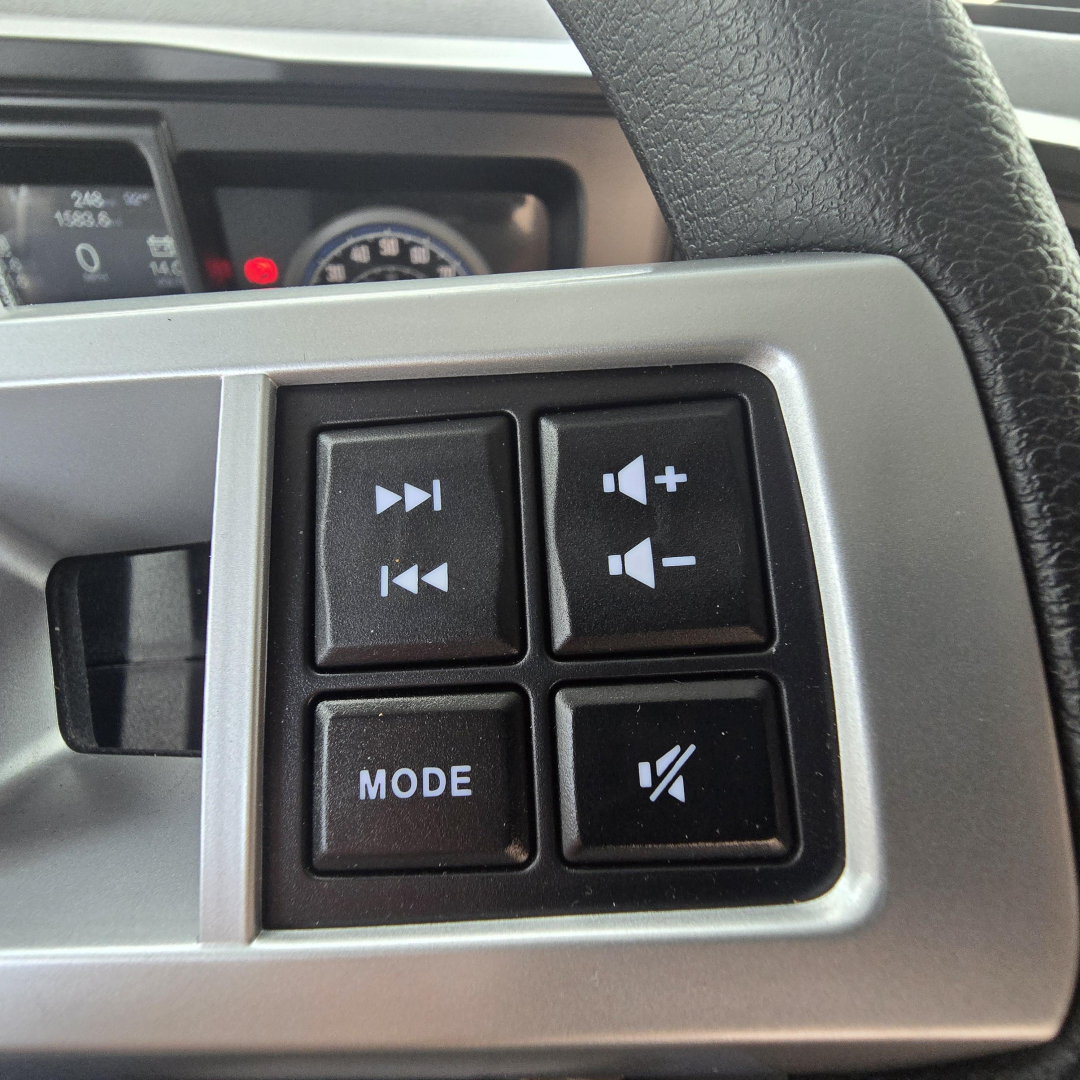Dashboard steering wheel controls in a Mack PI64T truck showing volume, mode, and mute buttons with a portion of instrument cluster in the background.