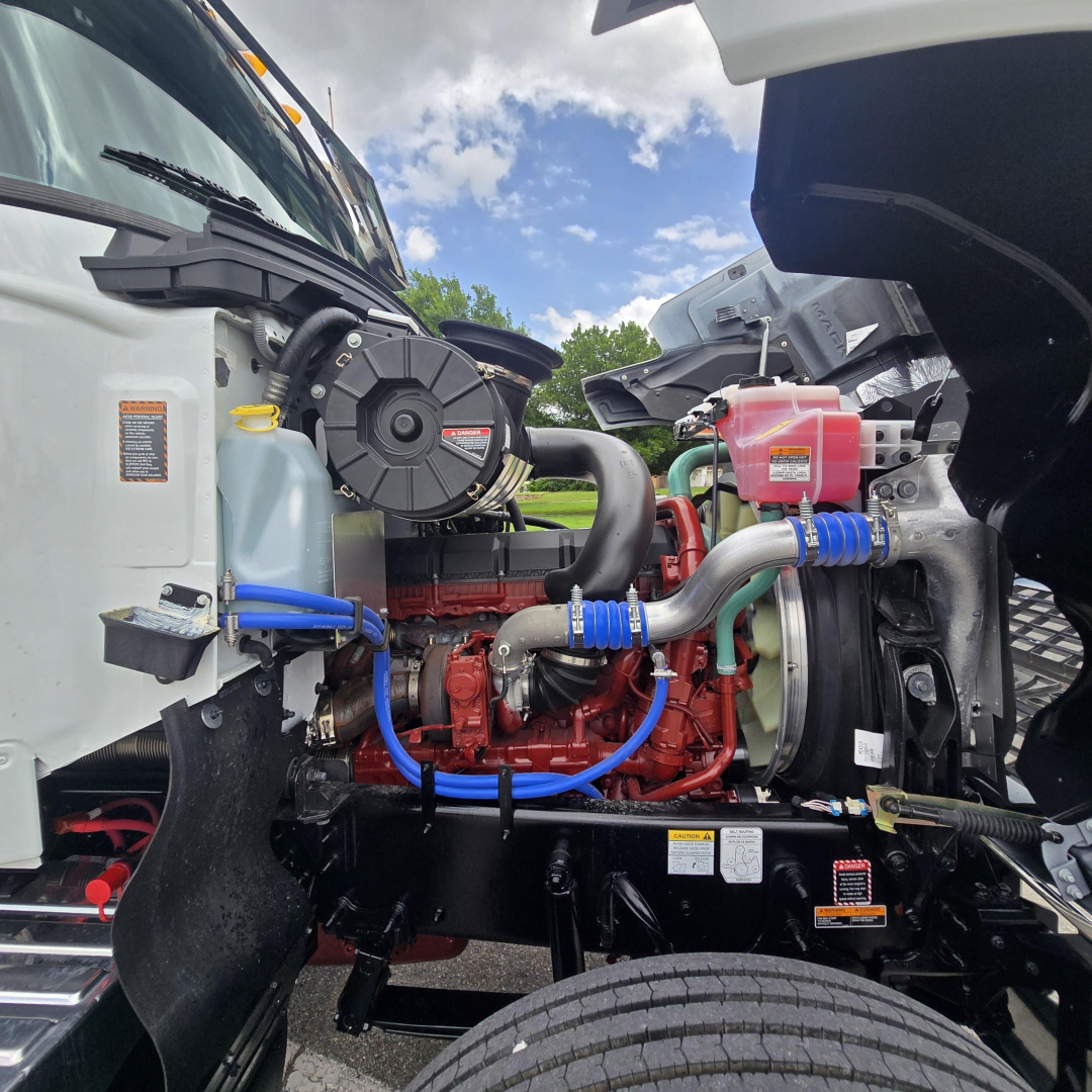 Mack PI64T engine bay showing diesel engine, blue hoses, coolant reservoir, air intake, and radiator at left side.