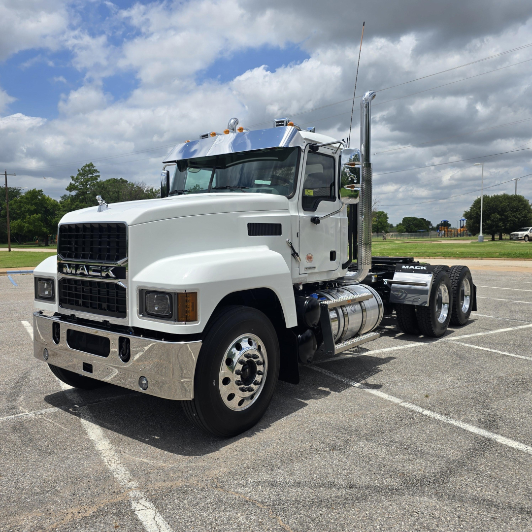 Mack PI64T conventional daycab truck in a parking lot, white cab with chrome bumper, dual rear axles, fuel tanks, and exhaust stack visible.