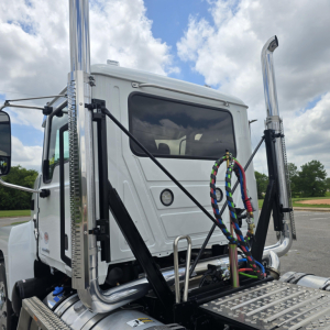 Front view of a 2027 Mack PI64T conventional daycab truck with chrome dual exhaust stacks, polished steps, and a customized wiring harness in the cab area.