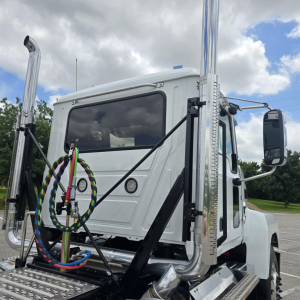 Truck cab and chrome exhaust stacks of a white 2027 Mack PI64T conventional daycab showing dual chrome air intake stacks and front windshield details.