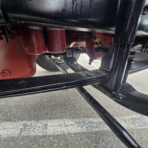 Underbody view of a new Mack PI64T conventional daycab showing chassis and suspension components, red engine block area, black frame rails, and exhaust pipe.