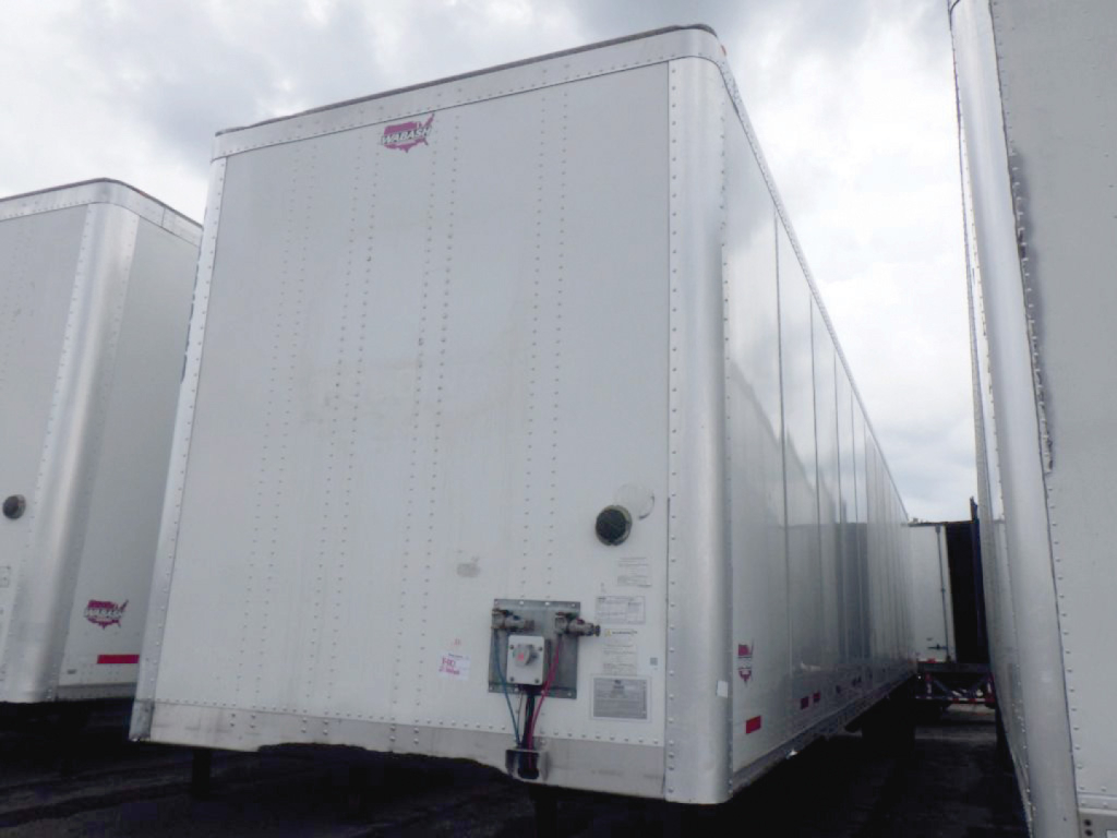 White box trailer showing front corner with small logo on top, door and utilities panel mounted near front. Text on label panels visible but not readable