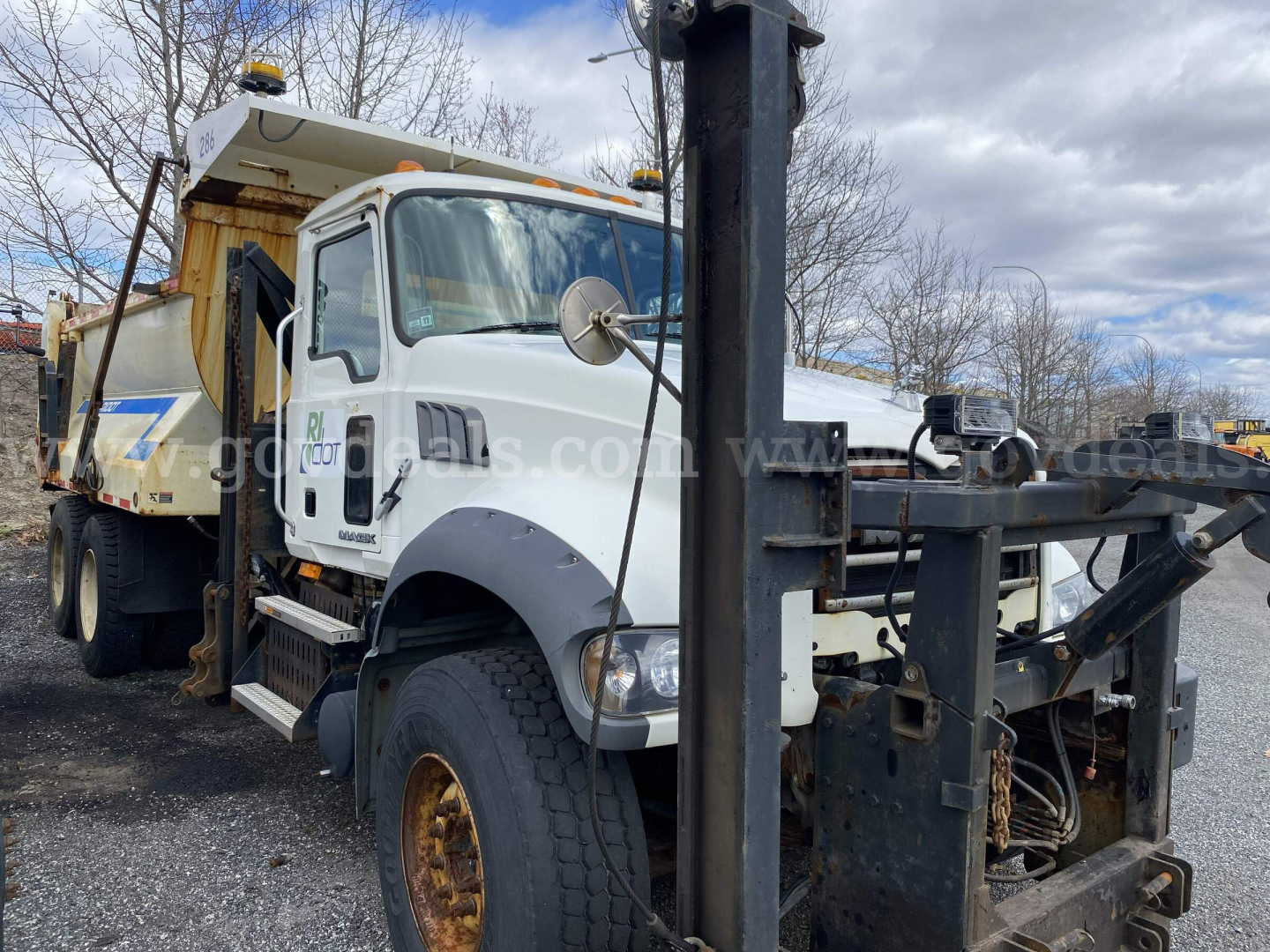 Dump truck front-left view of a white 2010 Mack GU713 with a mounted dump bed and crane, showing side mirror, steering wheel, and hydraulic components.