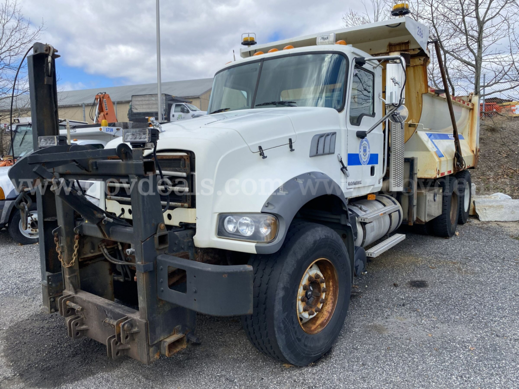 Mack GU713 dump truck in-service with front snow plow attachment and dump bed; white cab with blue emblem, stacked tires, and equipment on the frame.