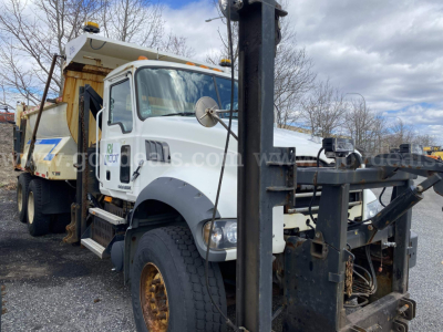 Dump truck front-left view of a white 2010 Mack GU713 with a mounted dump bed and crane, showing side mirror, steering wheel, and hydraulic components.