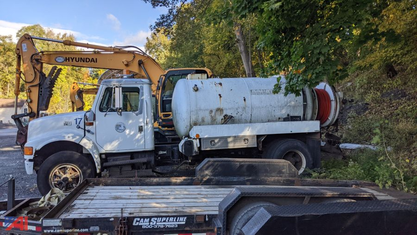 Side view of a white 1997 International 4900 sewer jet truck with a mounted large cylindrical tank and a Hyundai hydraulic excavator arm in the background.