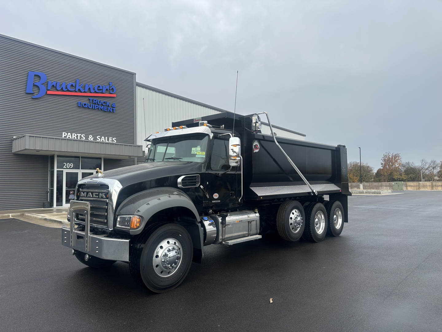 Dump truck showing Mack GR84F cab and dump bed, black exterior, chrome bumper, and dual rear axles in front of a parts shop.