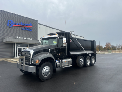 Dump truck showing Mack GR84F cab and dump bed, black exterior, chrome bumper, and dual rear axles in front of a parts shop.