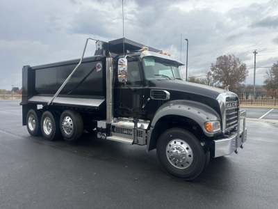 Dump truck with black cab and bed showing Mack grille, chrome mirrors, dual rear wheels, and fuel/tank area. Visible text includes Mack brand on grille and side badges, no other legible labels.