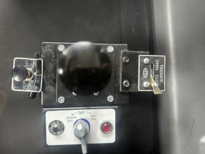Dump truck cab control panel with levers and labels showing OFF windup tarps and wind tarp controls, in-service panel with toggle switch and indicators.