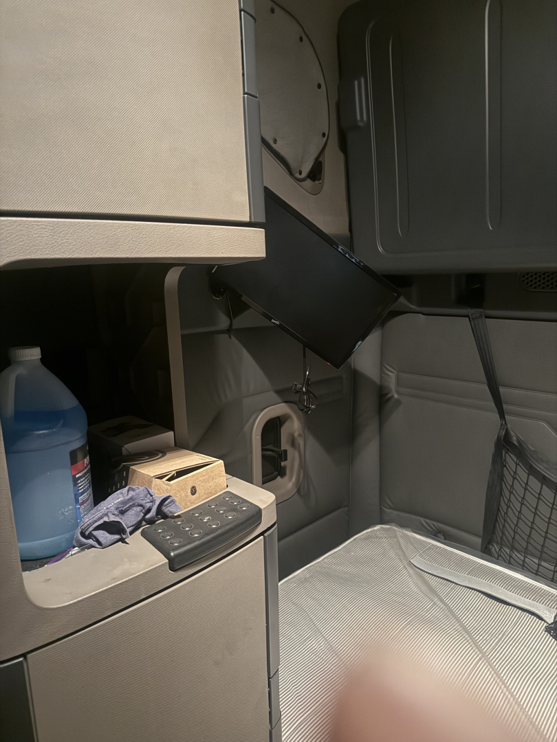 Truck cab interior showing sleeper area and storage compartments with a blue liquid container, a keypad, and a partially open cabinet.