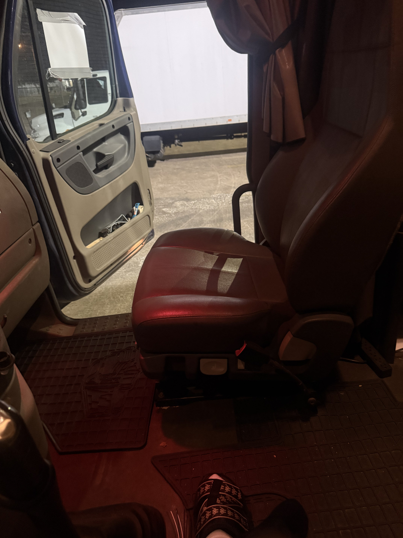 Seat and open cab door of a Freightliner Cascadia truck cabin, passenger seat visible with red upholstery, interior floor mats, exterior side mirror and doorway in view.