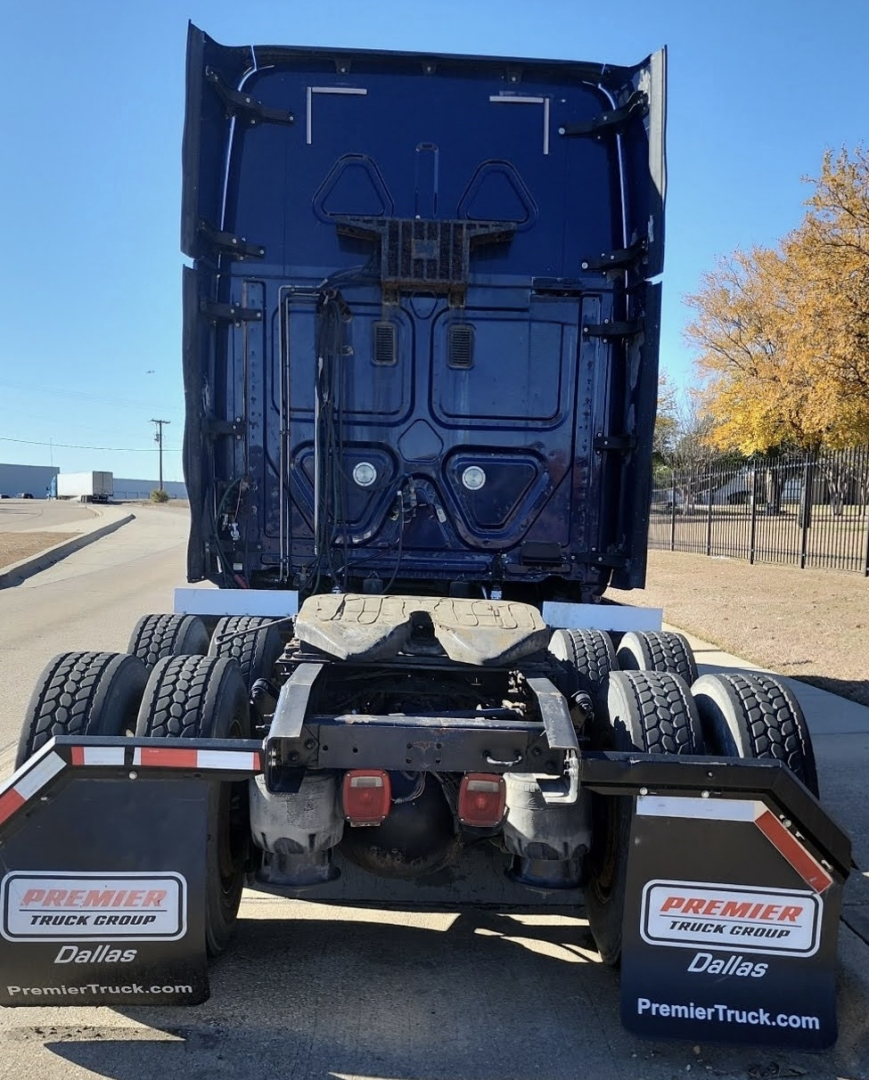 Freightliner Cascadia tractor rear view showing fifth wheel, dual rear axles, mud flaps with Premier Truck Group Dallas branding, blue cab underside and rear door area.