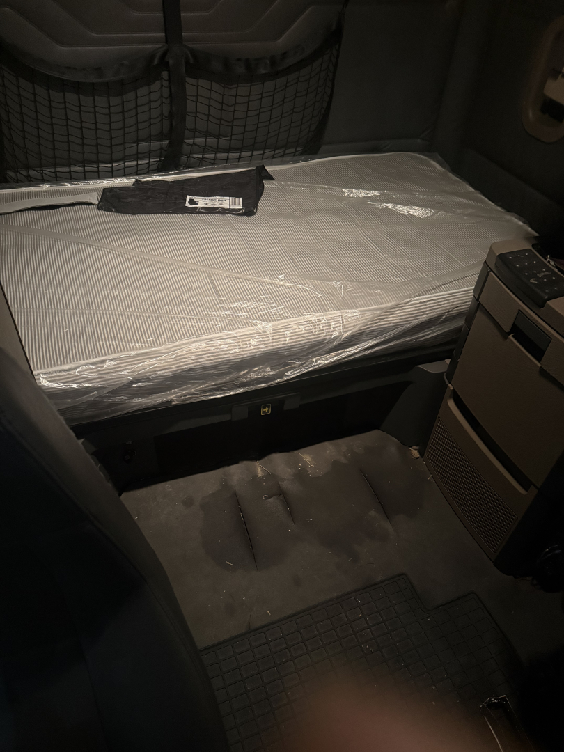 Freightliner Cascadia sleeper cab interior showing a wrapped mattress and storage cabinet with a side drawer and floor mat.