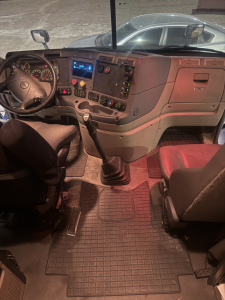 Truck cab interior of a 2015 Freightliner Cascadia conventional sleeper showing steering wheel, dashboard with gauges, gear shifter, and dual front seats.
