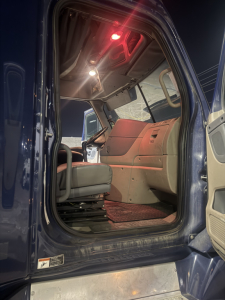 Cab interior of a Freightliner Cascadia truck viewed from the doorway showing the driver's seat and step area; steering wheel, dashboard, and side window visible, with ambient cabin lighting.
