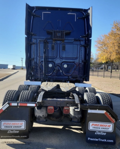 Freightliner Cascadia tractor rear view showing fifth wheel, dual rear axles, mud flaps with Premier Truck Group Dallas branding, blue cab underside and rear door area.