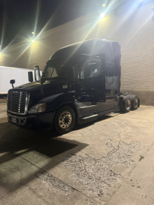 Truck tractor silhouette at night showing long hood, cab, and dual rear axles. Freightliner Cascadia cab with sleeper, black paint, headlight detail, and chrome wheel hubs.