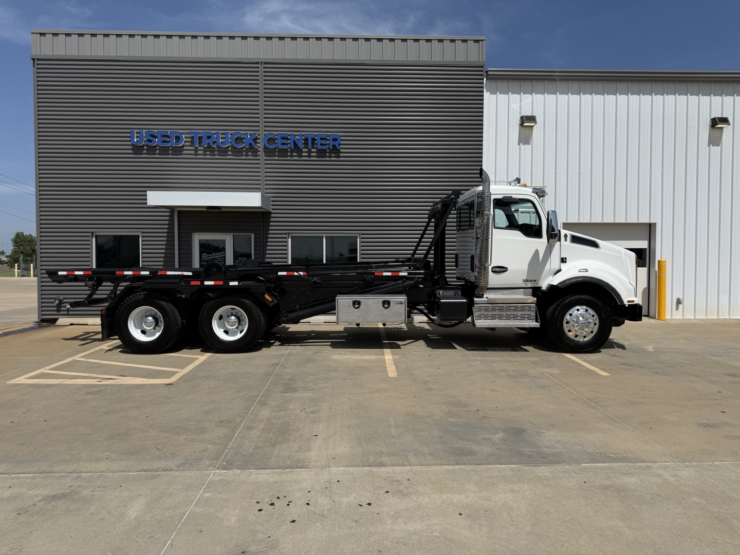 Kenworth T880 rolloff truck in front of dealership building with 'Used Truck Center' sign, white cab, black flatbed, dual rear axles, stock number B3204P, VIN 1NKZLP0X4JJ223978.