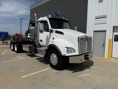 Kenworth T880 roll-off truck white cab with flatbed in a dealership lot, front grille and bumper visible.