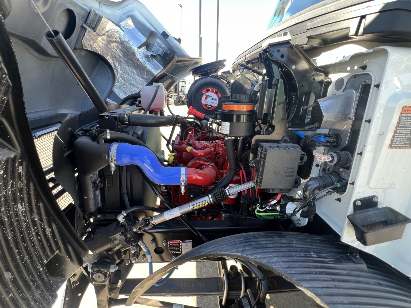 Mack MD7 engine bay showing red gasoline engine, blue coolant hose, black air intake, multiple hoses, and various fluids bottles under the hood.