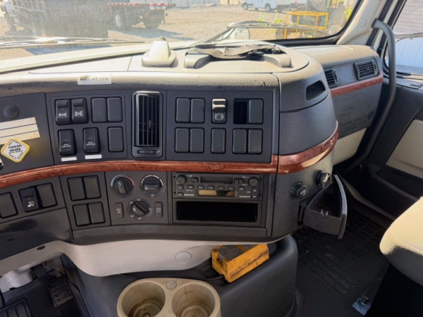 Cab interior of a Volvo VHD truck showing dashboard with numerous buttons, sliders, and gauges, wood grain trim, steering wheel on right side, cup holders visible.