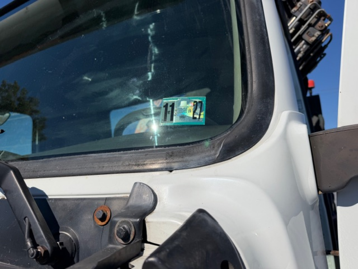 Truck cab window with a blue/green inspection sticker showing numbers '11' and '22' and a windshield in view; part of a crane truck in the background.