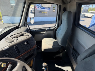 Volvo VHD truck interior cabin view showing driver's seat and dashboard with steering wheel and center console. Visible external signage through window reading 'USED TRUCKS' and other dealership posters.