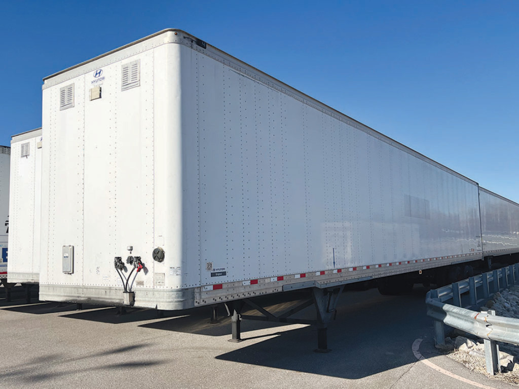 Tri-axle aluminum trailer with rear doors and dual wheels, white exterior, side fuel/electrical access panels, and landing gear visible at front.