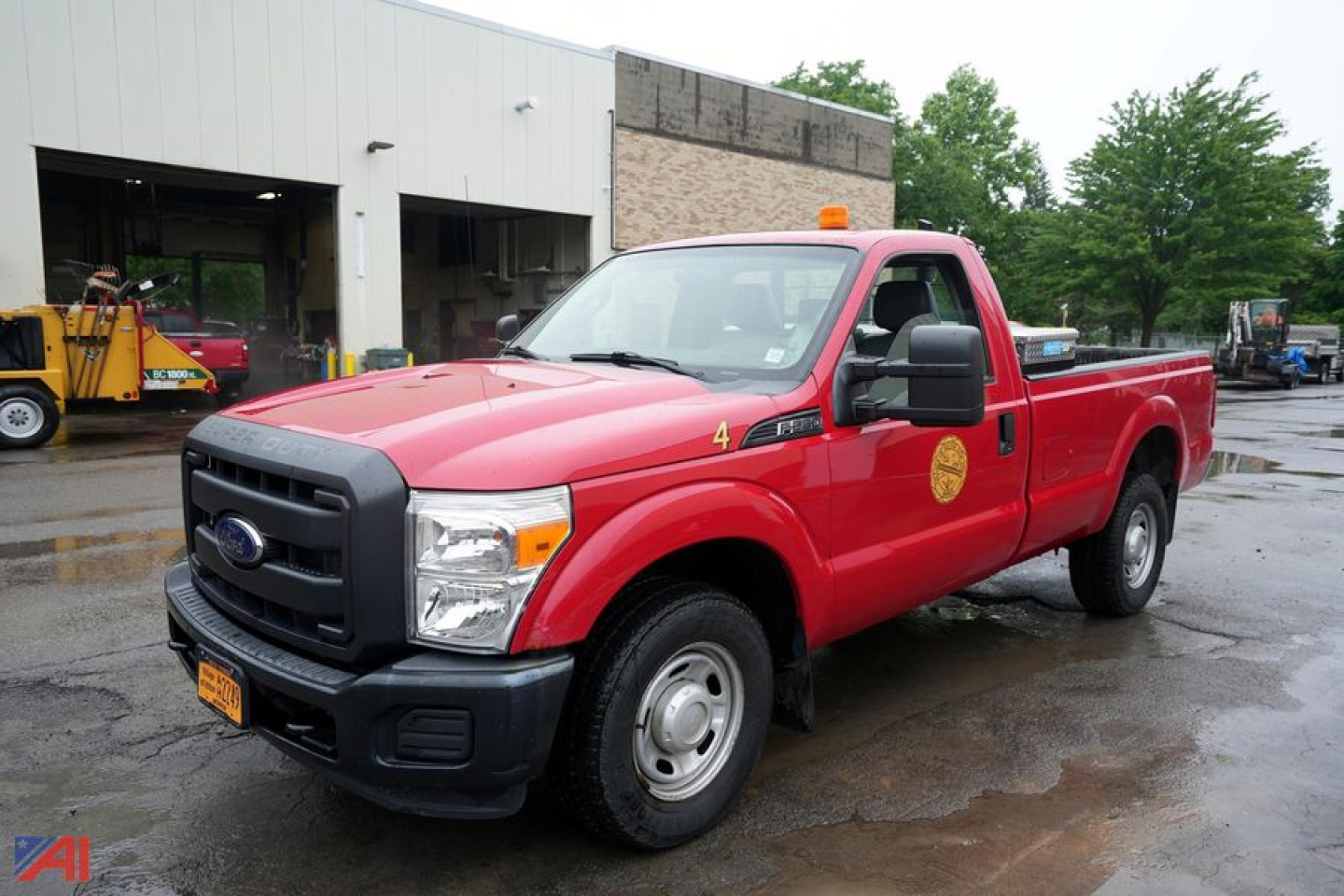 Red Ford F-250 XL single cab long bed pickup truck with orange beacon light on roof and toolbox in bed, front grille and bumper, dealership or service bay background.