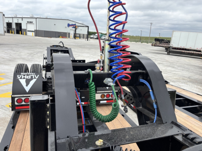 Trailer dolly chassis with blue and red coiled air hoses, green electrical line, and black steel frame. Visible are dual axles and wooden deck, with a purple/blue coil and hydraulic components.