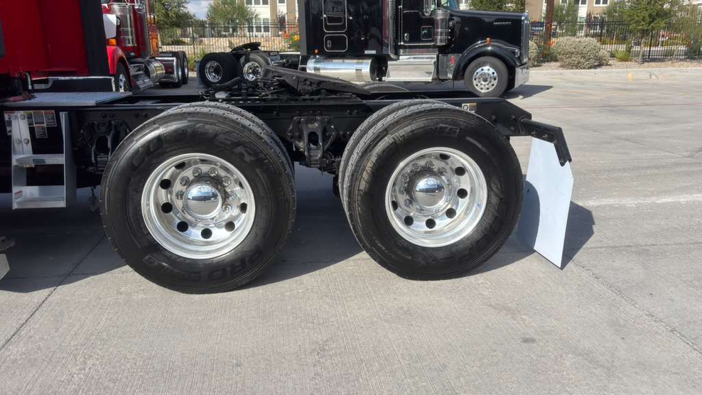 Kenworth T680 chassis side view showing dual rear wheels with chrome hubs and fender, concrete ground, other trucks in background.