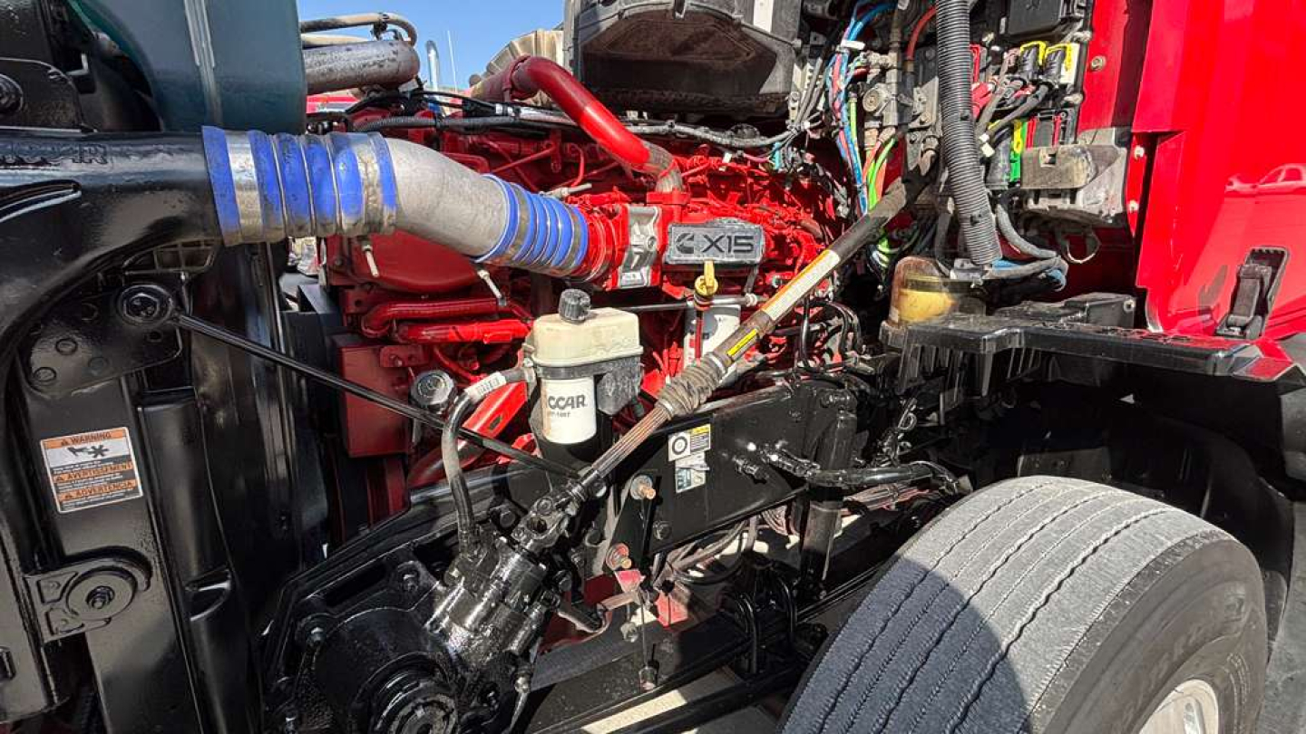 Truck engine bay with red engine block and blue hose clamps, various labels visible including white reservoir with 'COOL' or 'COOL' text, black frame, cables, and metal components.