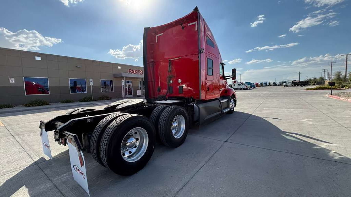 Kenworth T680 tractor-trailer in red, side view showing rear wheels and sleeper cab, numbers and dealership lot in background.