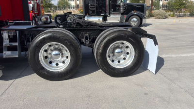 Kenworth T680 chassis side view showing dual rear wheels with chrome hubs and fender, concrete ground, other trucks in background.