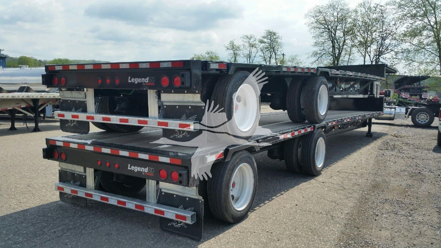 Manac drop deck trailer, side view showing tandem axle with dual wheels and storage platforms; visible labels include 'Legend' on the rear bumper; flatbed rails with reflective tape.