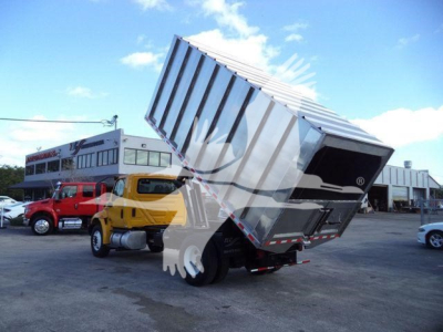 Truck with tipping dump bed raised in a yard, International 4300 chassis, yellow cab, chipper equipment in the back, dealership buildings in background.