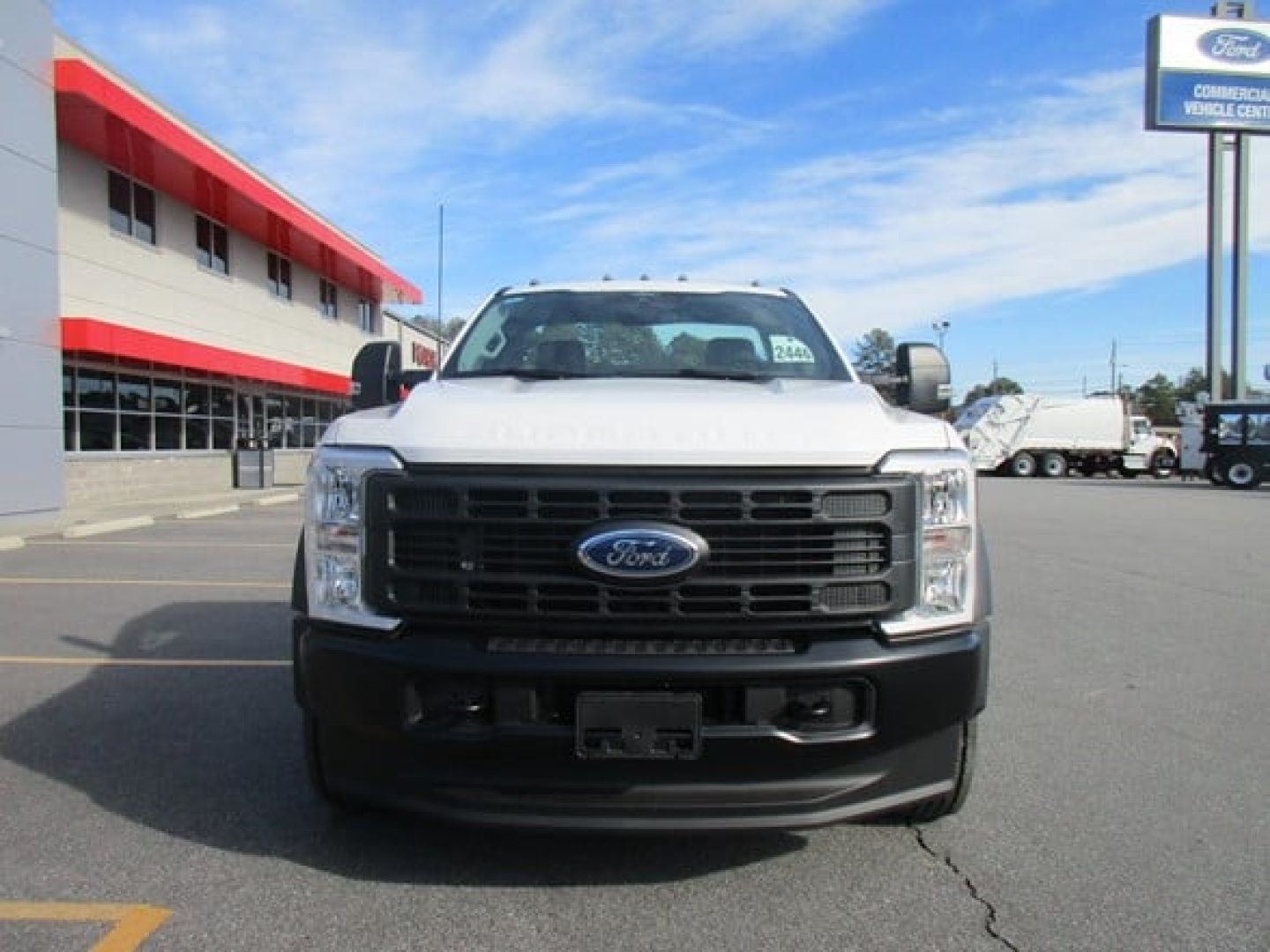 Ford F-450 cab and chassis front view showing large black grille with Ford emblem, white body, parking lot, dealership building in background.