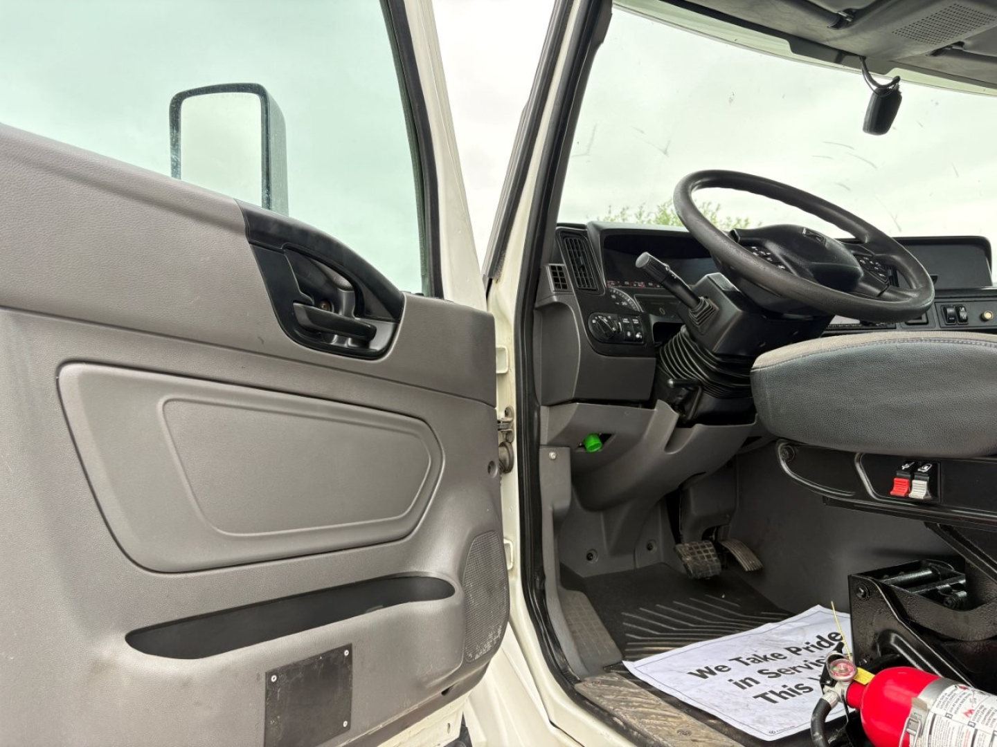 Truck cab interior showing driver's door open, steering wheel and dashboard; some text visible on paperwork on floor mat near pedal.