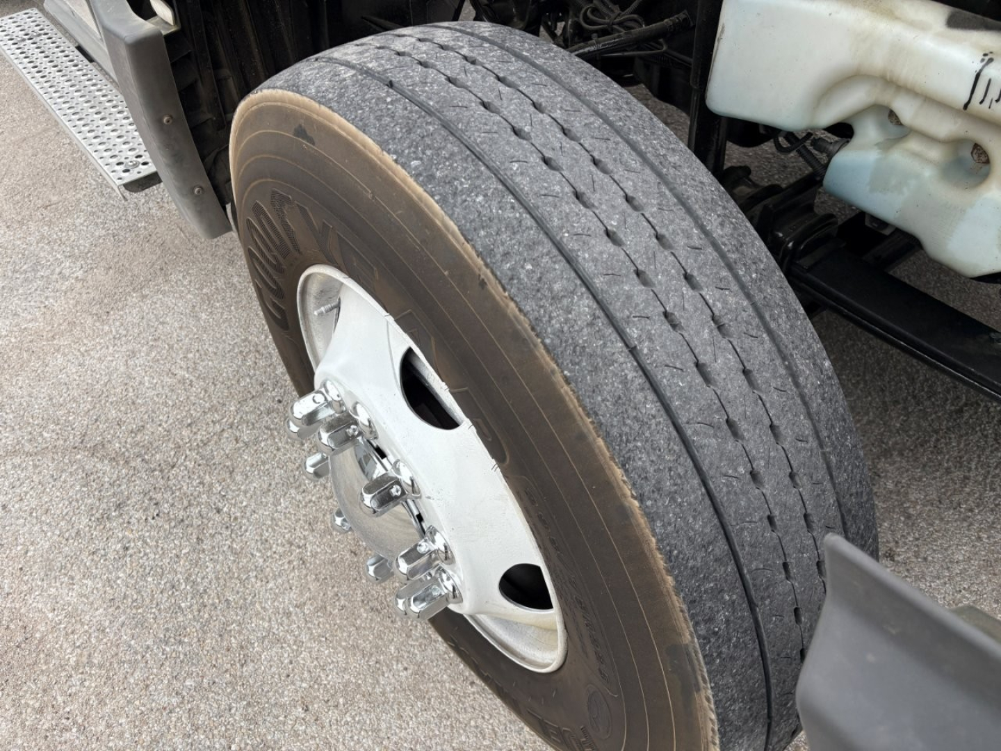 Truck wheel and front tire close-up showing hub and lug nuts with a portion of the truck frame and coolant reservoir nearby.