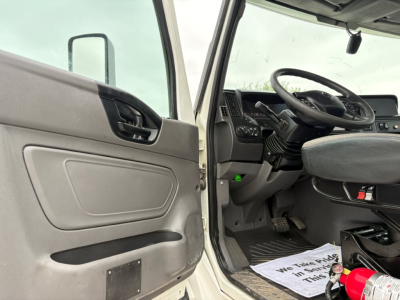 Truck cab interior showing driver's door open, steering wheel and dashboard; some text visible on paperwork on floor mat near pedal.