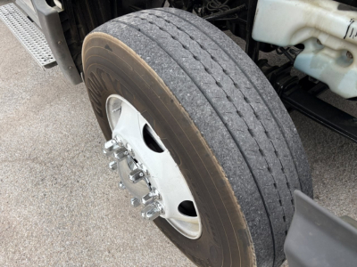 Truck wheel and front tire close-up showing hub and lug nuts with a portion of the truck frame and coolant reservoir nearby.