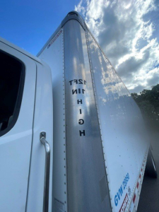 Box van truck cargo trailer with reflective metal side and HINO lettering on vertical seam; white cab visible; clear sky background.