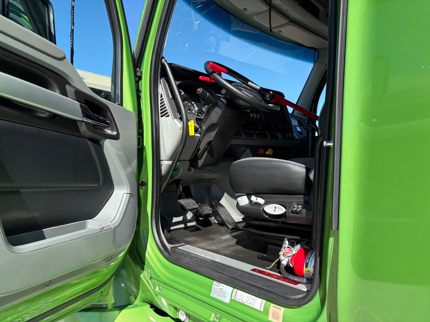 Kenworth T680 cab interior view from open driver door showing steering wheel, dashboard, seat, and pedals.