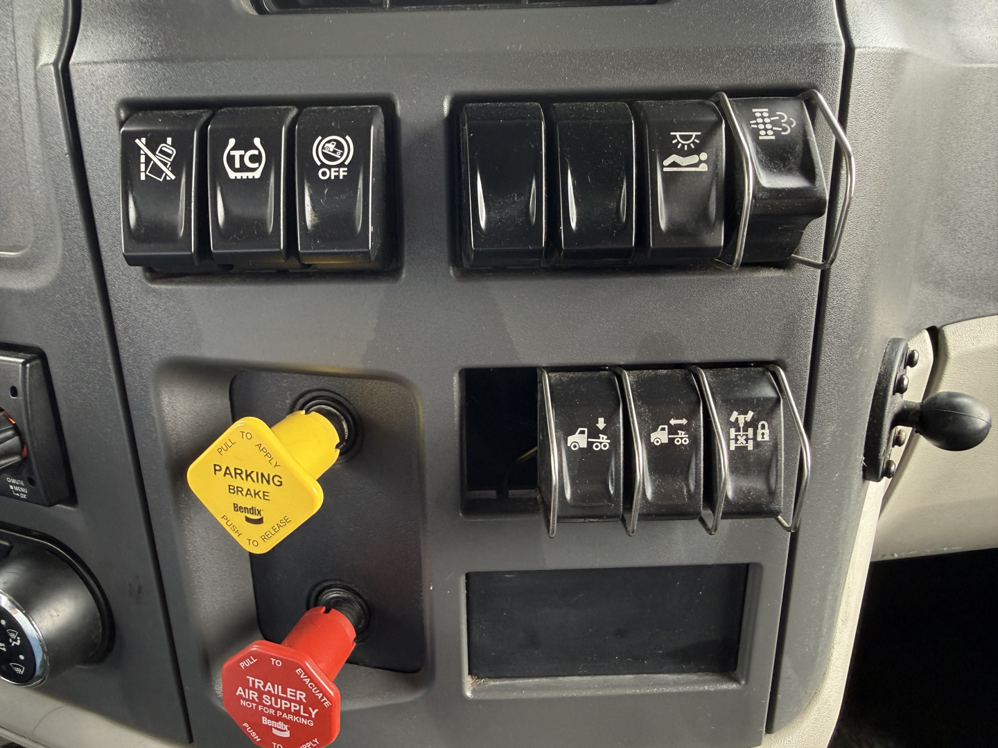 Peterbilt 579 dash cluster showing multiple switch panels and yellow parking brake knob; visible stickers read parking brake and trailer air supply; on the left a red trailer air supply knob; various icon buttons with OFF and other symbols.