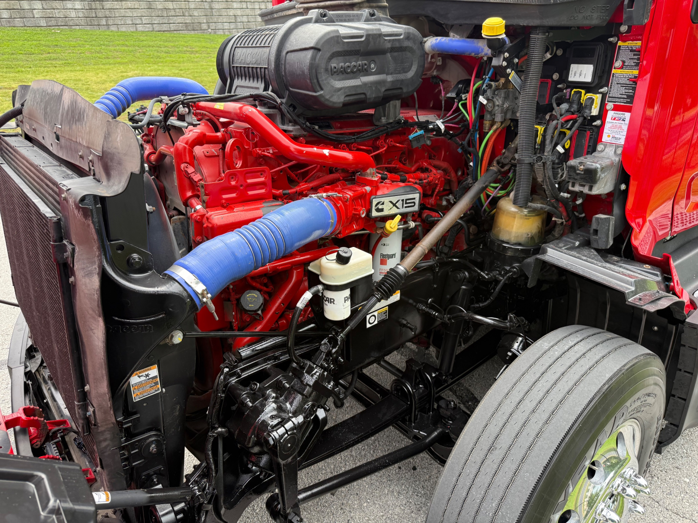 Peterbilt 579 engine bay showing red engine with blue turbo hoses, black radiator, and various wiring; blue air intake hose labeled EXIS, white reservoirs, and front tire visible.
